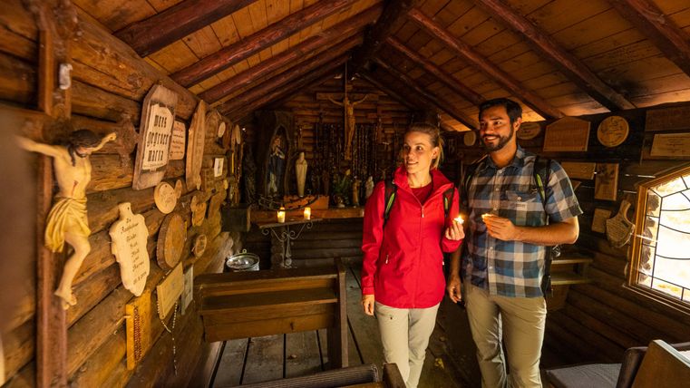 Ein gemütlicher Holzraum mit religiösen Figuren und Dekorationen. Zwei Personen stehen lächelnd und erkunden die Umgebung.