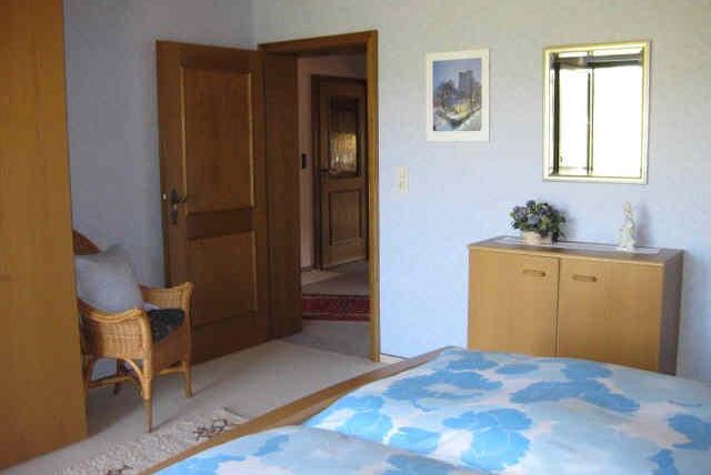 A cozy bedroom with a double bed and blue floral bedding. There is a wicker chair and a wooden dresser that complement the room.