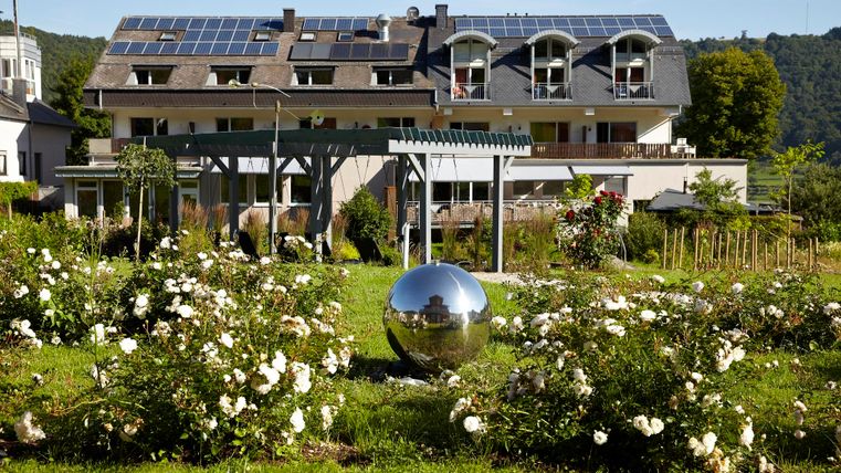 A modern building with solar panels stands in a beautiful garden. The garden is decorated with numerous roses and a large, shiny sphere.