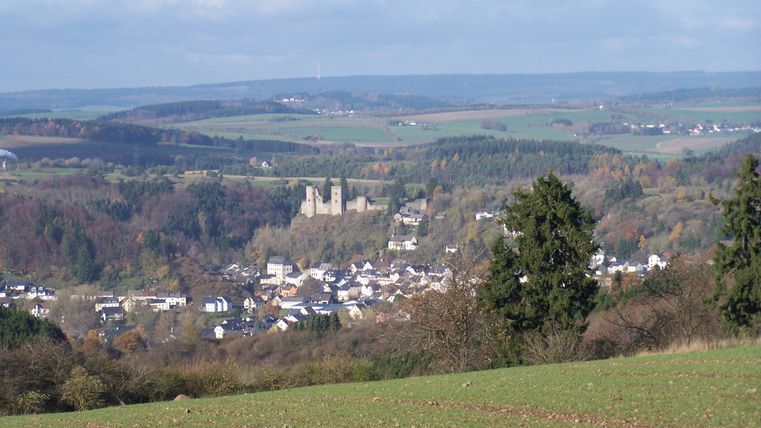 Eine malerische Landschaft mit sanften Hügeln und einer kleinen Stadt im Tal. Im Hintergrund ist eine Burg sichtbar, die über die Umgebung wacht.