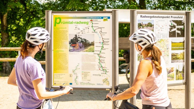 Twee fietsers staan voor een informatiebord over de Prüm-Vallei-fietsroute. Ze dragen helmen en bekijken de kaart en informatie.