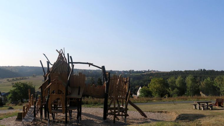 Eine kunstvolle Skulptur aus Holz ist im Freien zu sehen. Im Hintergrund erstreckt sich eine hügelige Landschaft unter einem klaren Himmel.