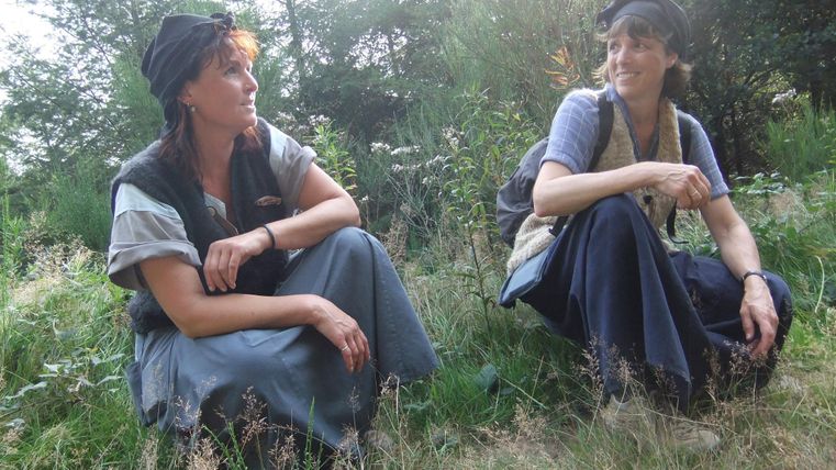 Two women are sitting on the ground in a green environment. They are wearing simple, traditional clothing and seem to be engaged in a conversation.