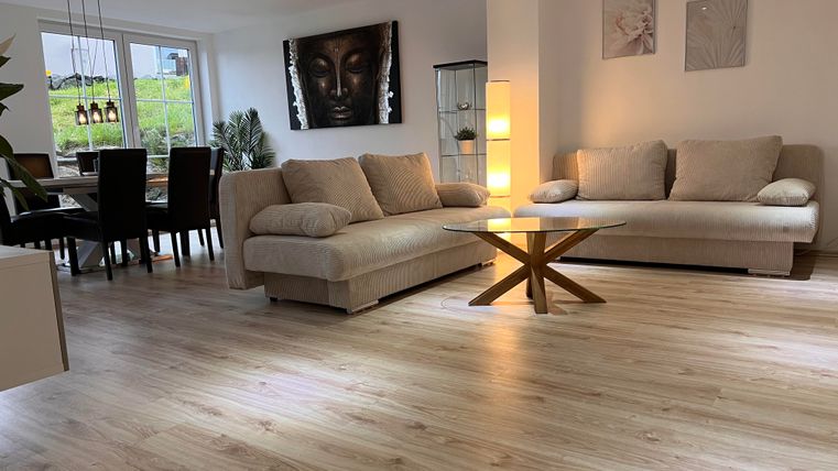 A modern living room with light furniture and a large window. There is a cozy seating area and a dining table in the background.