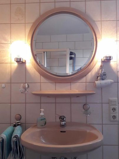 Een badkamer met een ronde spiegel, een wastafel en twee wandlampen. Er zijn handdoeken en verzorgingsproducten op een plank.