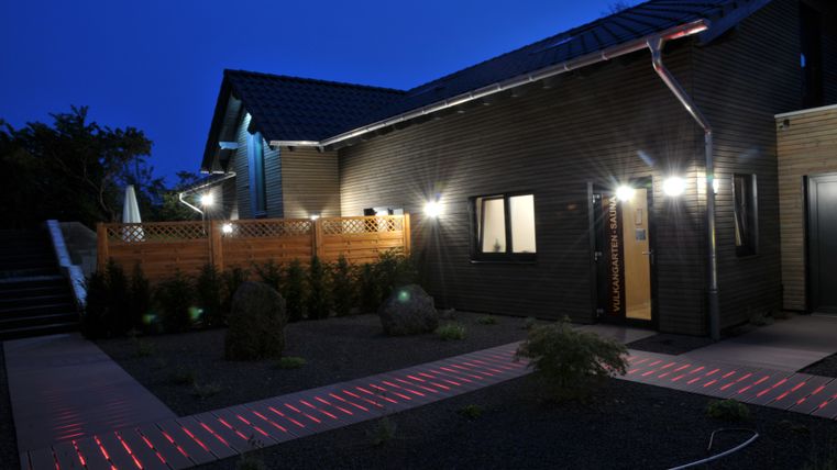 Ein modernes Haus mit Holzfassade und beleuchtetem Eingangsbereich bei Nacht. Der Weg zum Haus ist stilvoll mit roten Lichtern markiert.