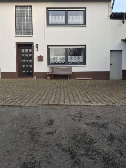 Ein zweigeschossiges Haus mit einem hellen Äußeren. Vor dem Haus befindet sich ein gepflasterter Bereich und eine Sitzbank.