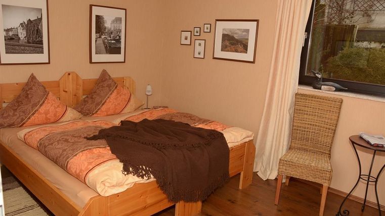 A cozy bedroom with a large bed and decorative pillows. Pictures hang on the walls, and a chair is beside the window.