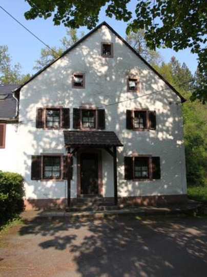 A white house with brown windows and a gabled roof. It stands surrounded by trees in a quiet environment.