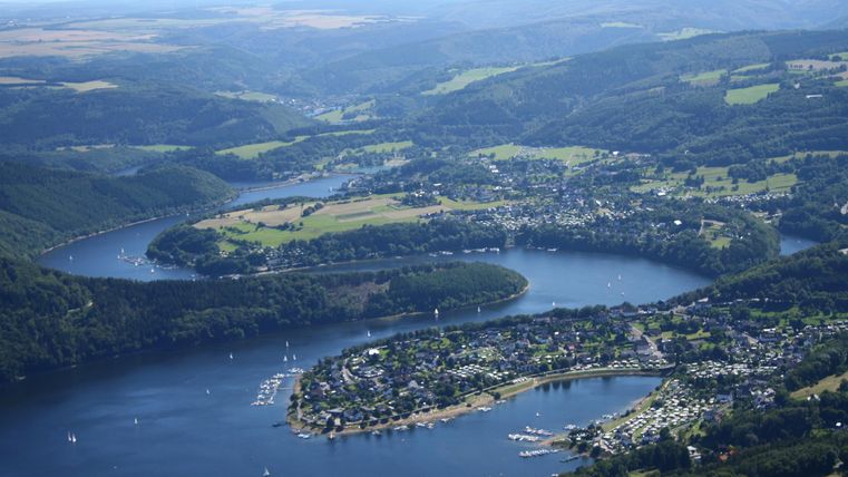 A picturesque landscape with a vast lake and surrounding hills. The green shore is lined with small towns and boats.