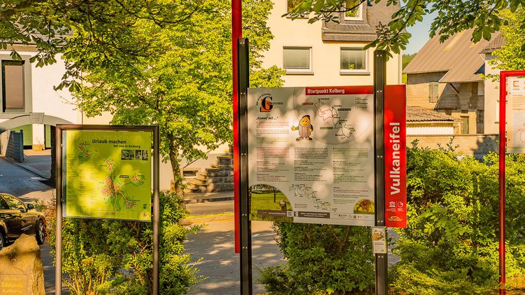 Een informatief bord staat tussen bomen en huizen. De omgeving lijkt groen en uitnodigend.