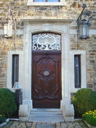 Eine elegante Holztür mit kunstvollem Beschlag, eingerahmt von steinernen Wänden. Auf beiden Seiten der Tür stehen grüne Buchsäulen.