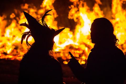 Twee silhouetten staan voor een groot vuur. Een persoon draagt een opvallende hoed, terwijl de ander spreekt.