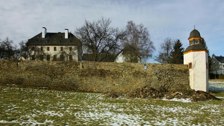 Oberehe Castle with an old stone wall and a small tower in the foreground. The ground is partly covered with snow, trees without leaves.