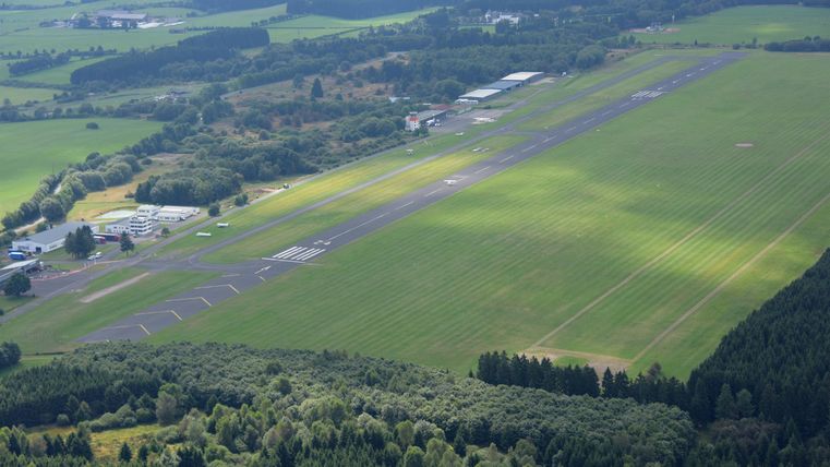 Een uitgestrekt vliegveld met groen gras en een blauwe lucht. Op de achtergrond zijn bomen en enkele gebouwen zichtbaar.