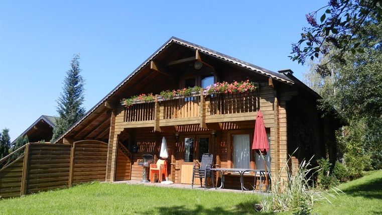 Ein hübsches Holzhaus mit einem Balkon voller Blumen. Der Garten ist grün, und im Hintergrund sieht man Bäume unter einem klaren blauen Himmel.