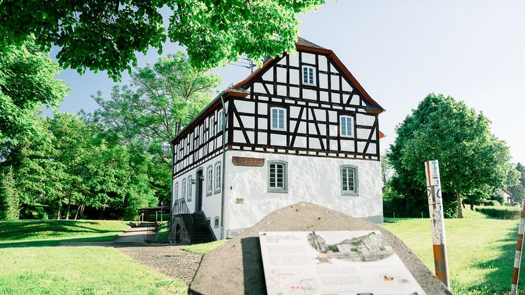 Een historisch vakwerkhuis omringd door bomen en groenvoorzieningen. Op de voorgrond is een informatieschild te zien.