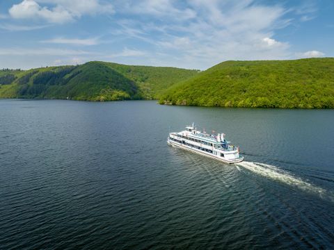 A ship glides calmly over a lake, surrounded by green hills. The sky is blue with some clouds.